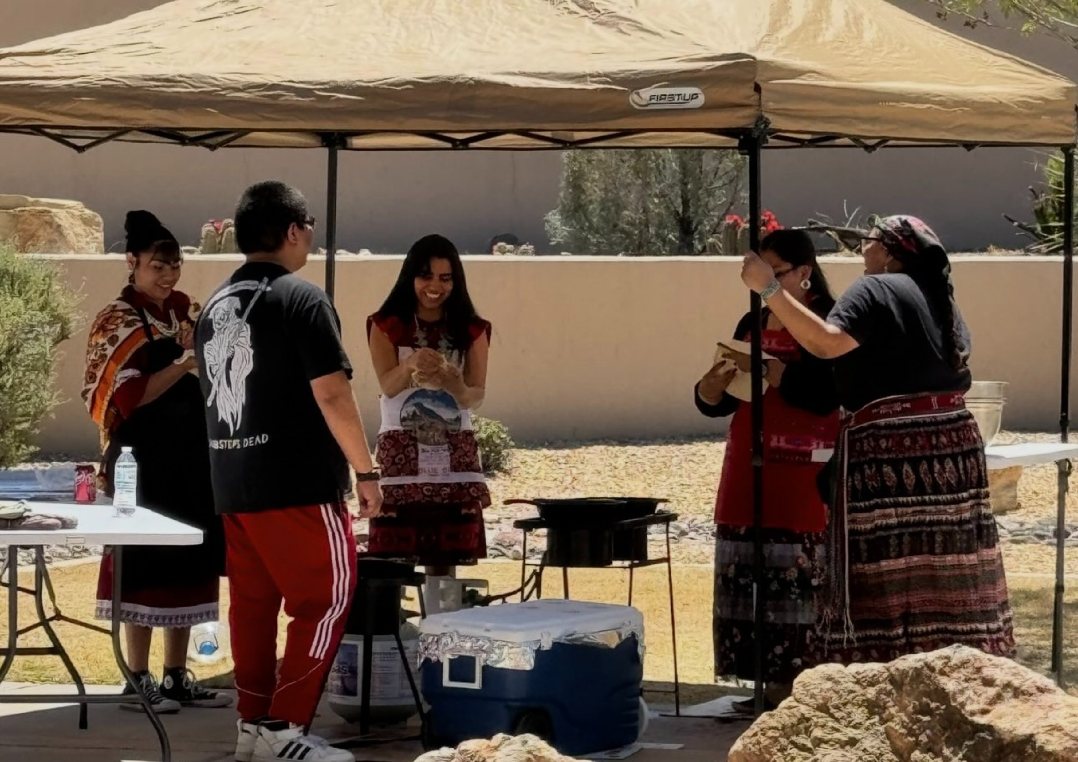 AIW Indian Taco Frybread Making. A group of four people, three women and one man, under a beige canopy tent in a sunny, desert-like setting.